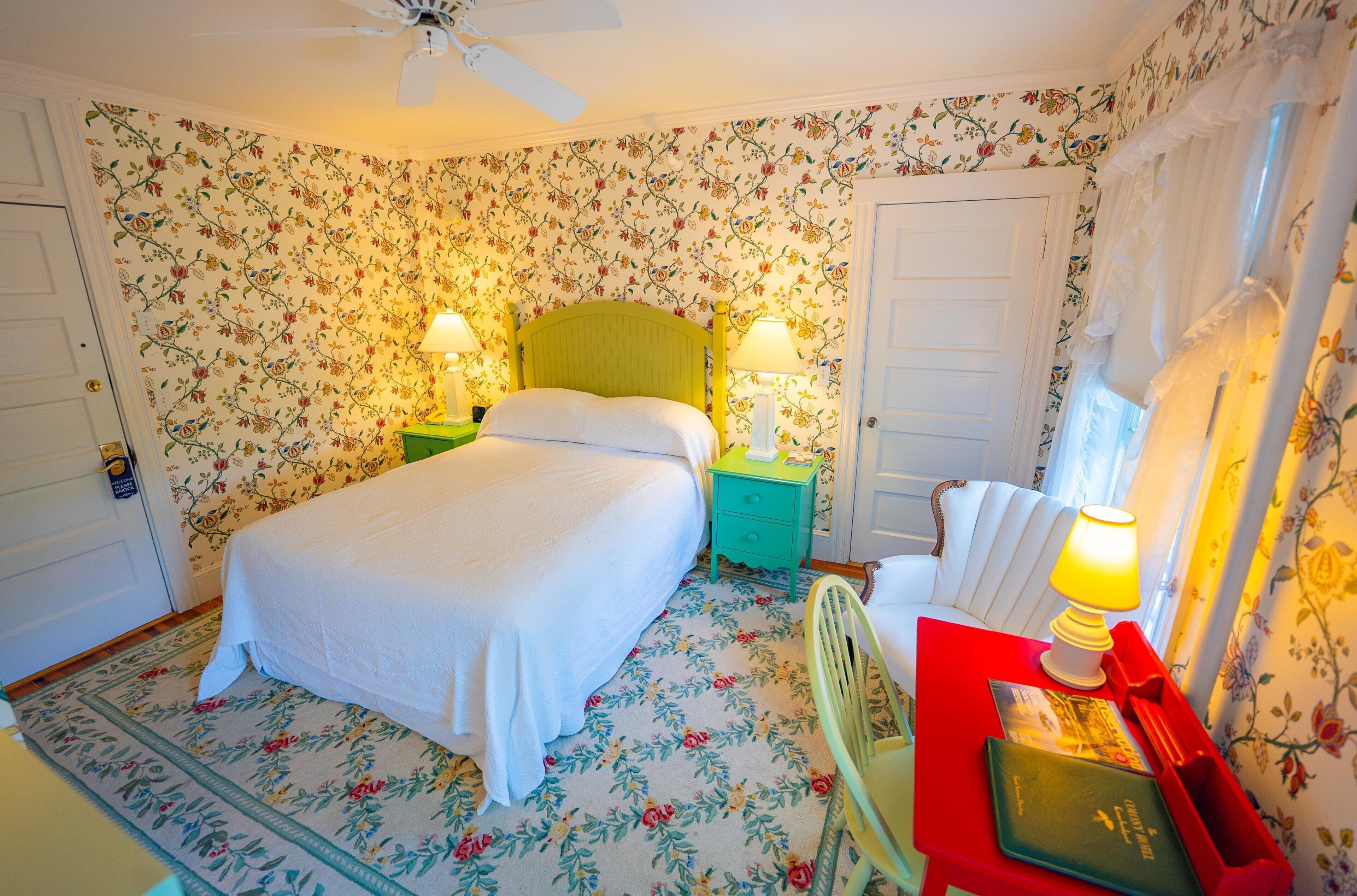 Een slaapkamer met bloemenbehang, een bed met witte lakens, twee nachtkastjes met lampen, een rode stoel en een rode tafel met een lamp.