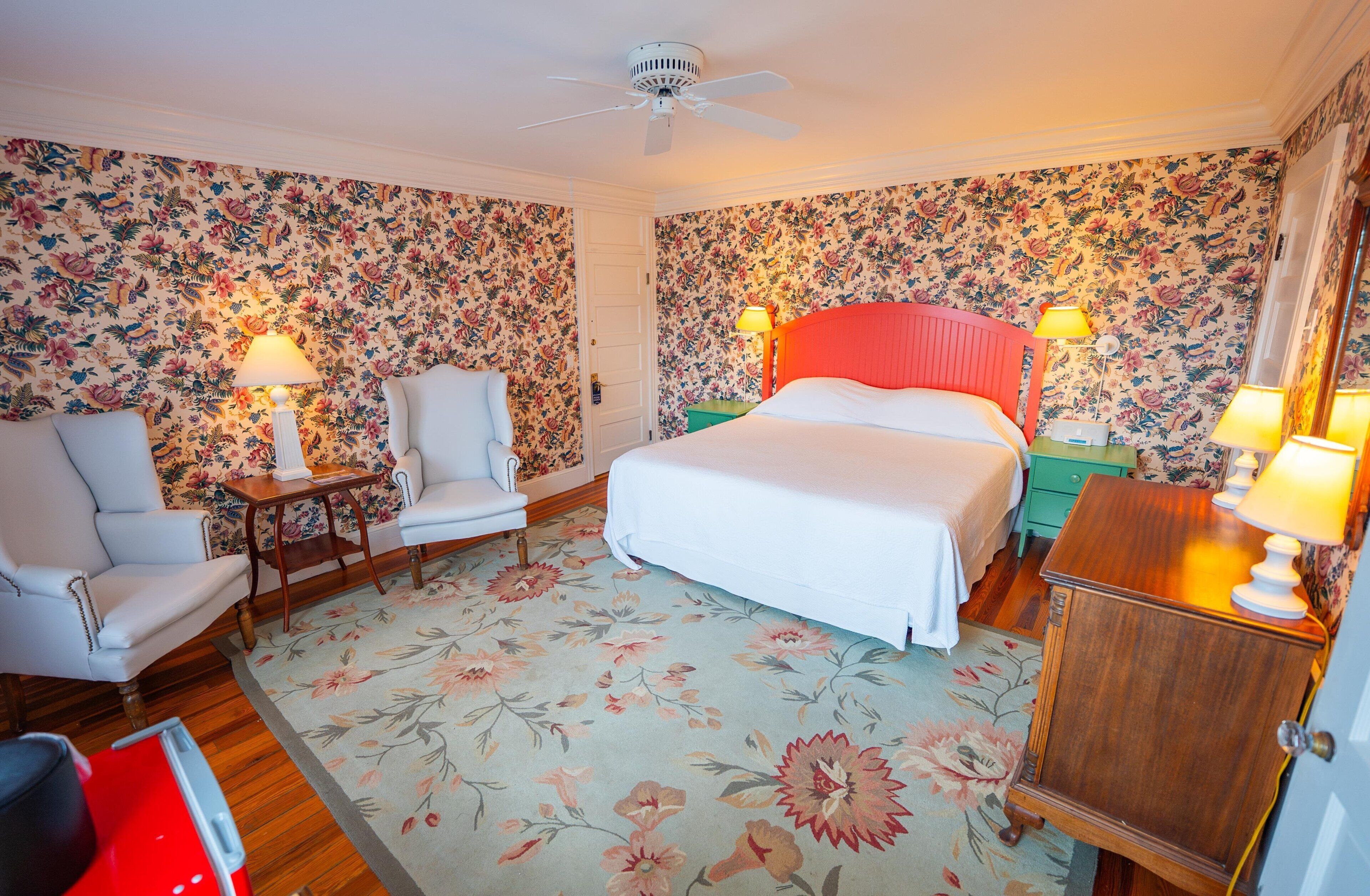 A hotel room with a bed, a chair, a desk, a nightstand, a wardrobe, and a ceiling fan.