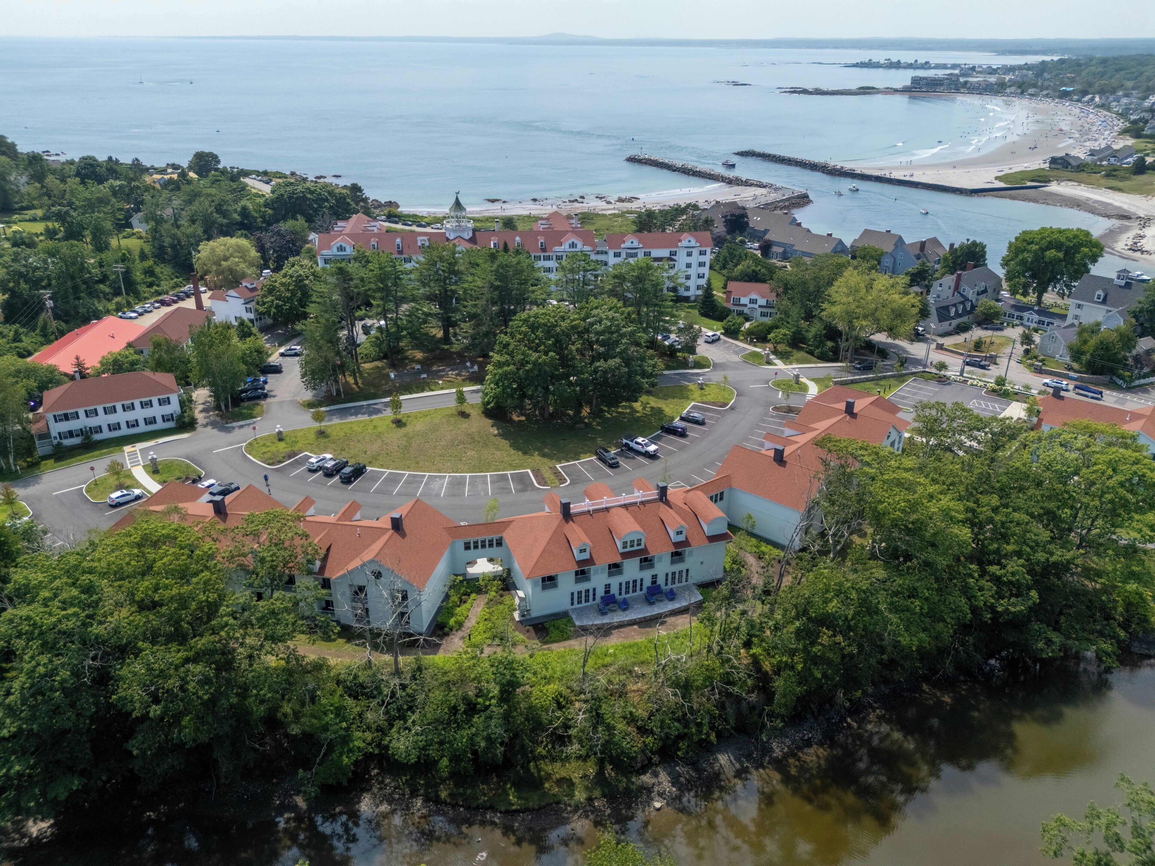 An aerial view of a coastal town with a central roundabout, buildings with red roofs, and a beach in the background.