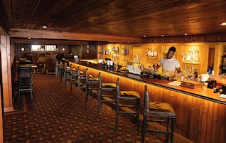 Área de bar com balcões de madeira, banquetas de couro e tapete estampado.