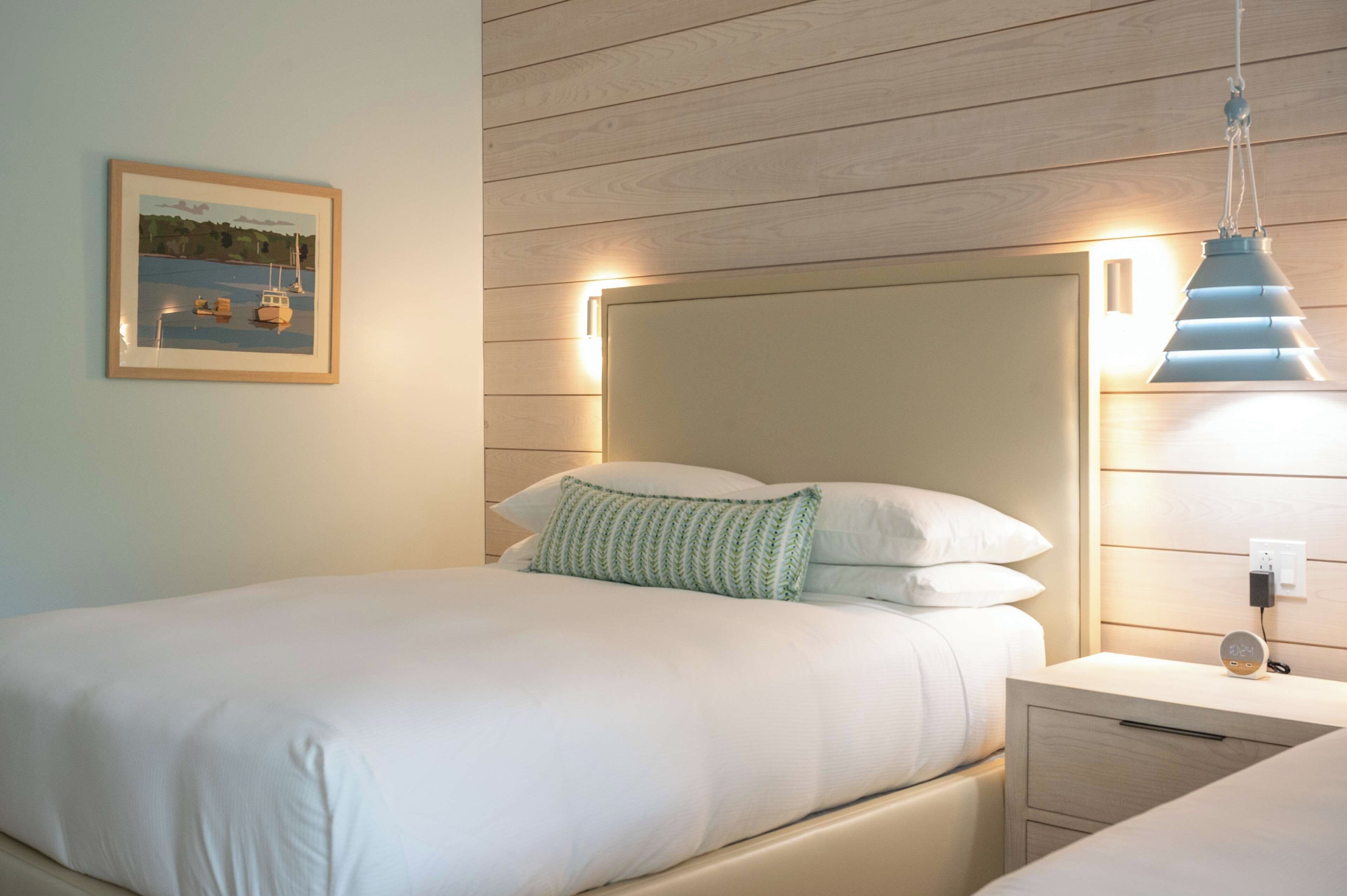 A neatly made bed with white linens and a single green pillow, a bedside table with a lamp, and a framed picture on the wall.