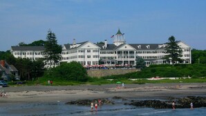 A large, multi-story hotel with a prominent tower and flag, situated on a beachfront with rocky areas and a sandy beach. - The Colony Hotel (Kennebunkport)