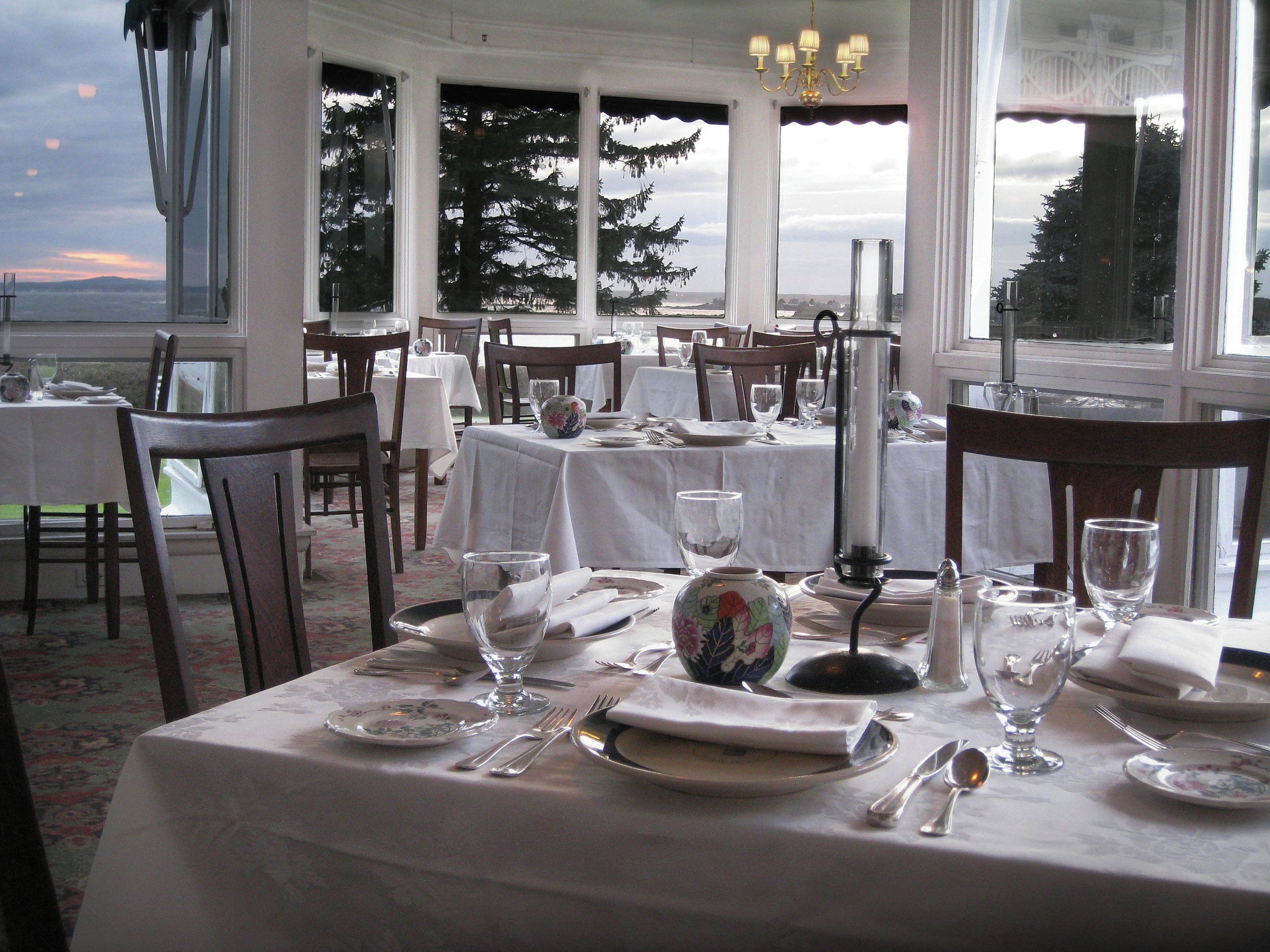 Un espace repas avec des tables dressées, de grandes fenêtres donnant sur la verdure et un lustre.