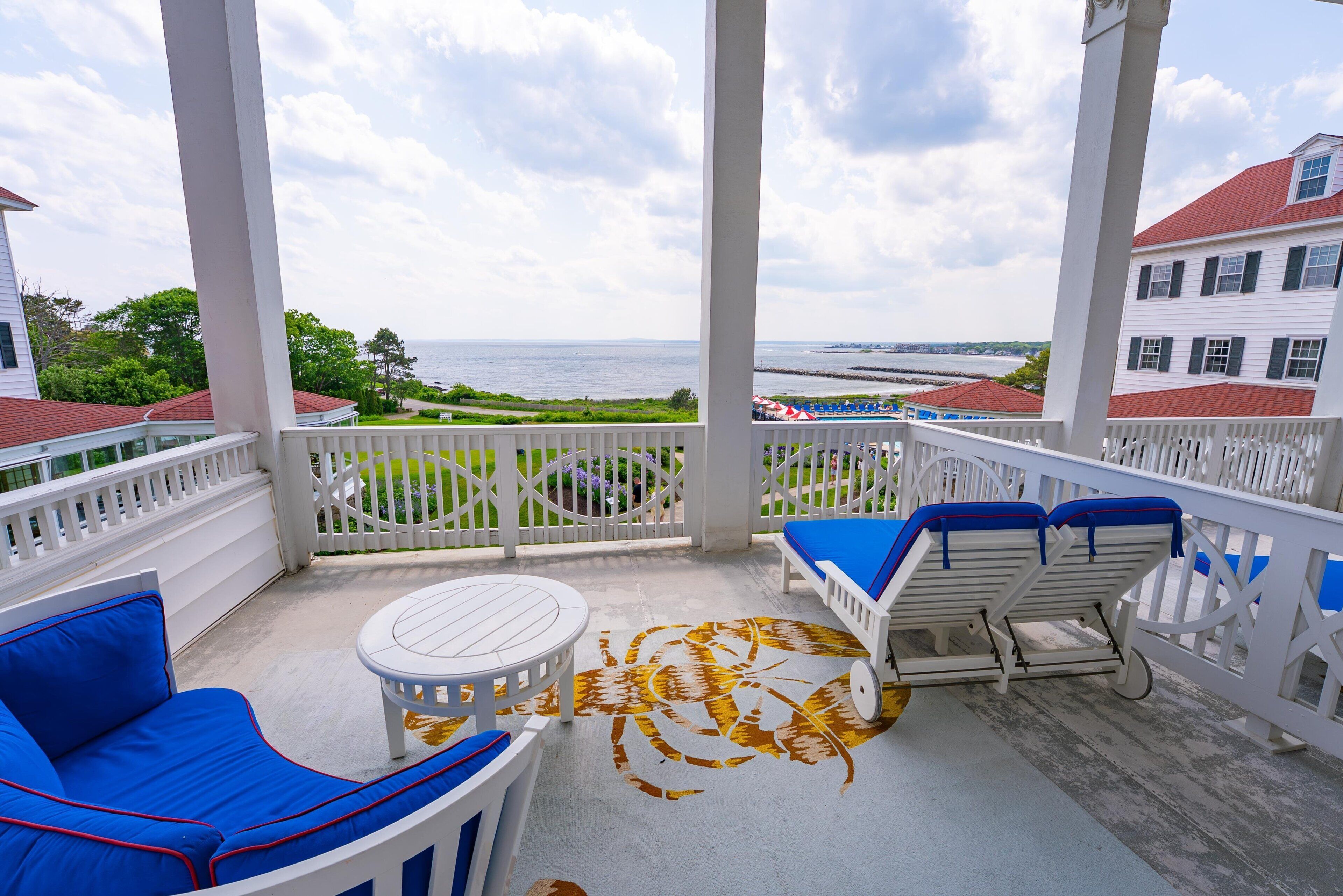 Un espace extérieur abrité, avec vue sur la mer, agrémenté d’une balustrade blanche, de sièges bleus rembourrés et d’une petite table ronde.