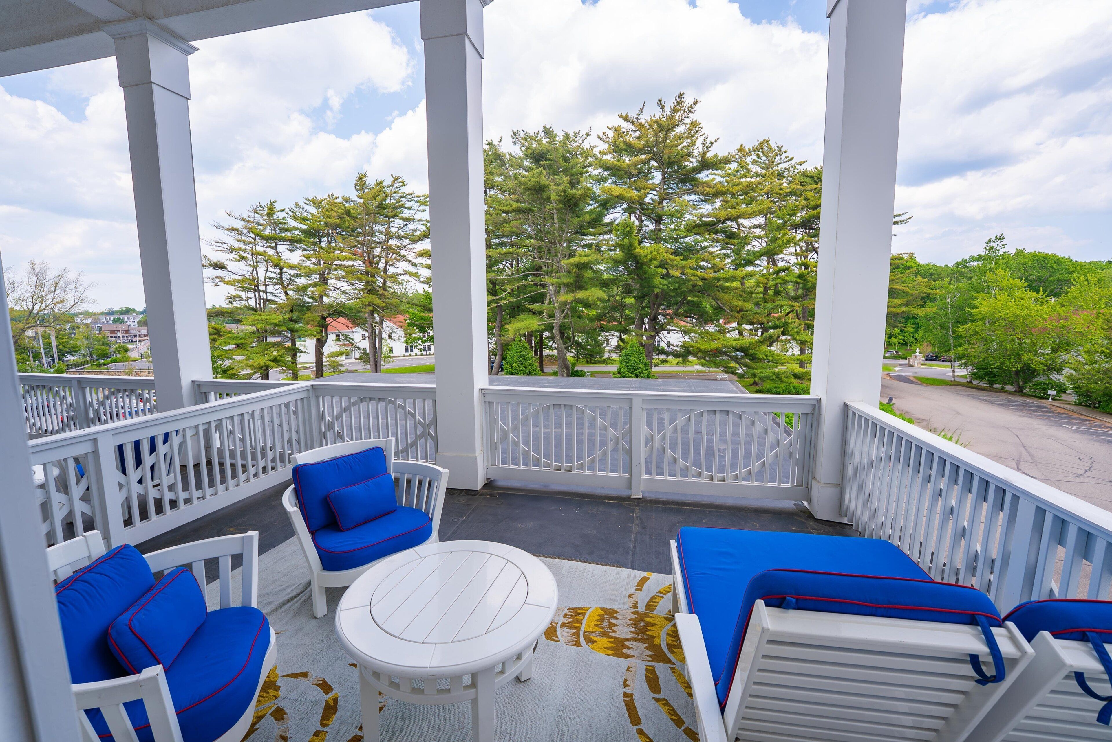 A covered outdoor seating area with white furniture and blue cushions, overlooking a residential street and trees.
