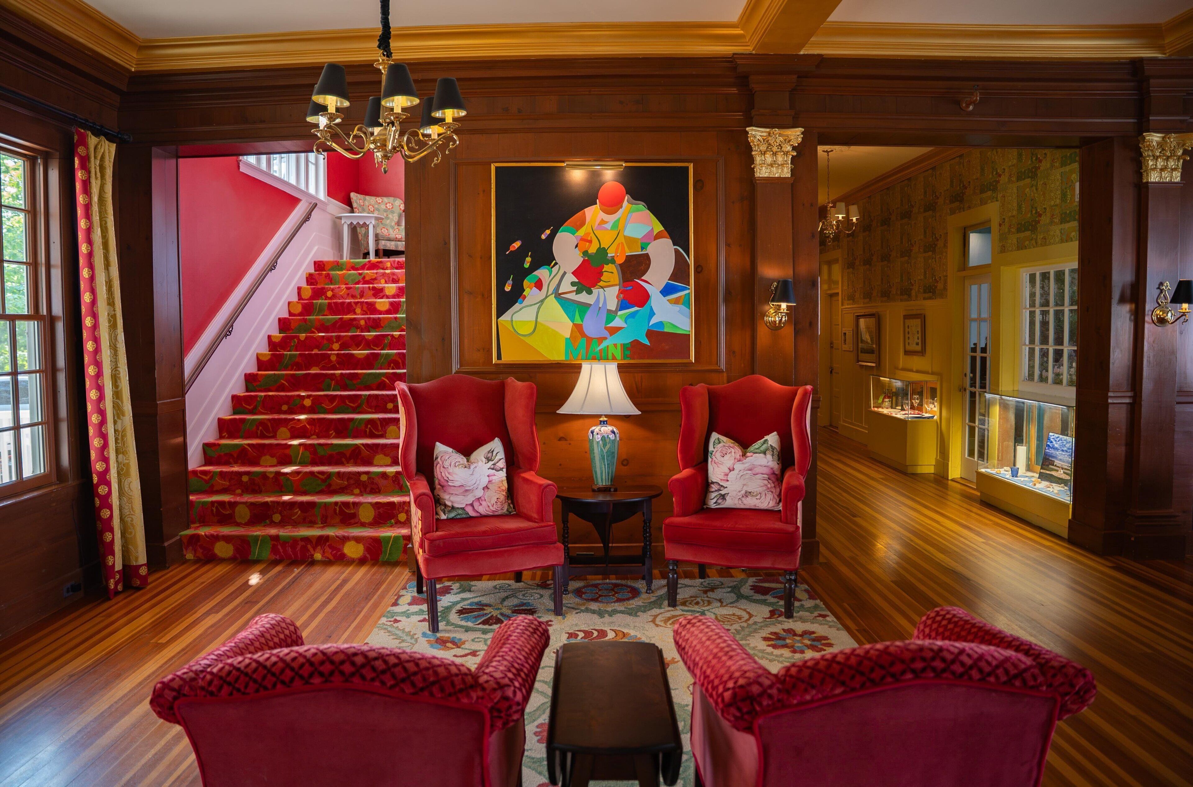 A grand staircase with red carpet, two red armchairs with floral cushions, a table with a lamp, and a colorful abstract painting on the wall.