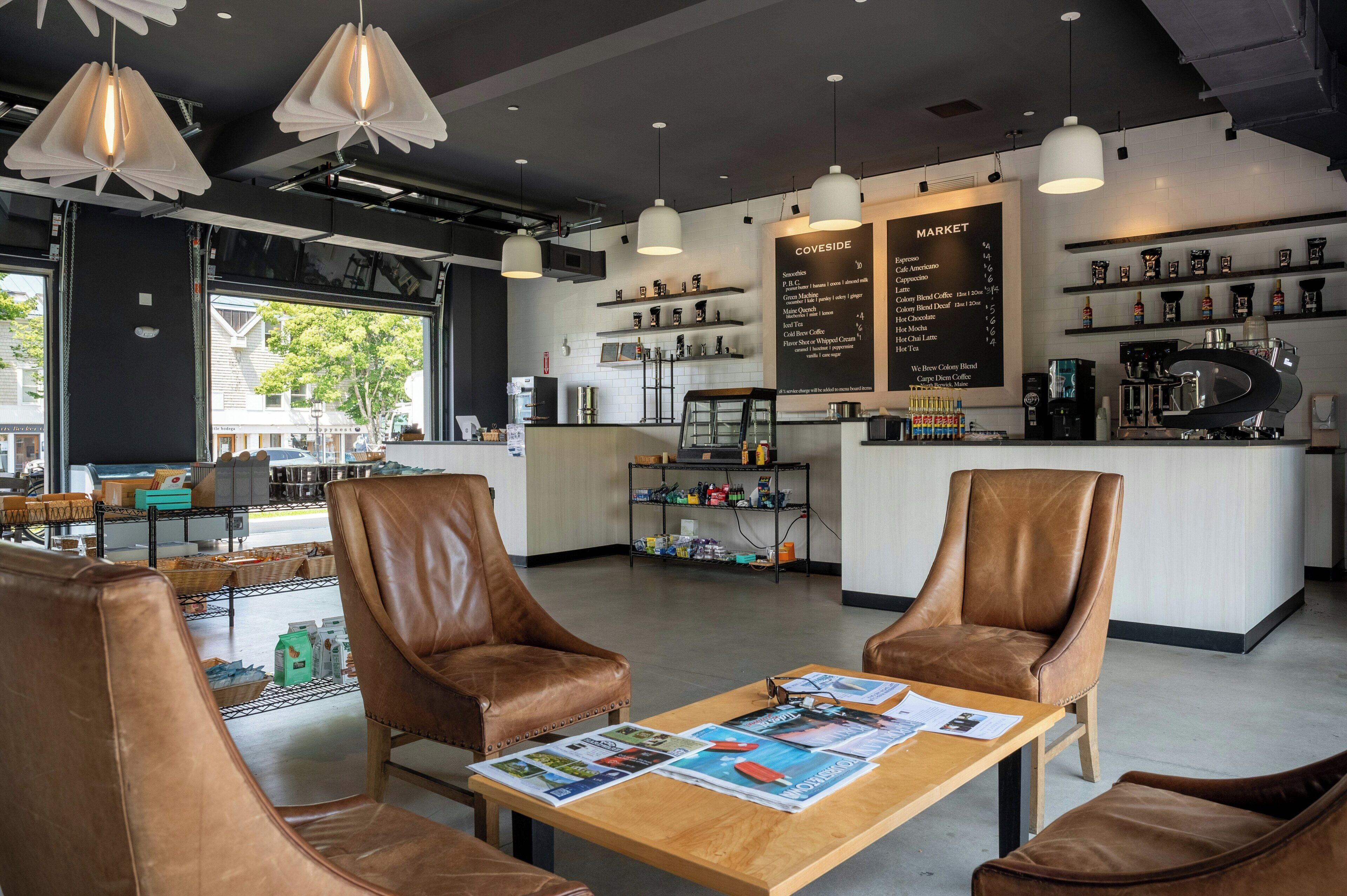 A modern cafe interior with leather chairs, a wooden table with magazines, a coffee machine, and a menu board.