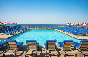 A poolside area with blue lounge chairs and red umbrellas, overlooking a clear blue sea.