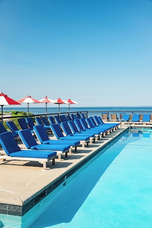 A poolside area with blue lounge chairs and red umbrellas, a clear sky, and a view of the ocean. - The Colony Hotel (Kennebunkport)
