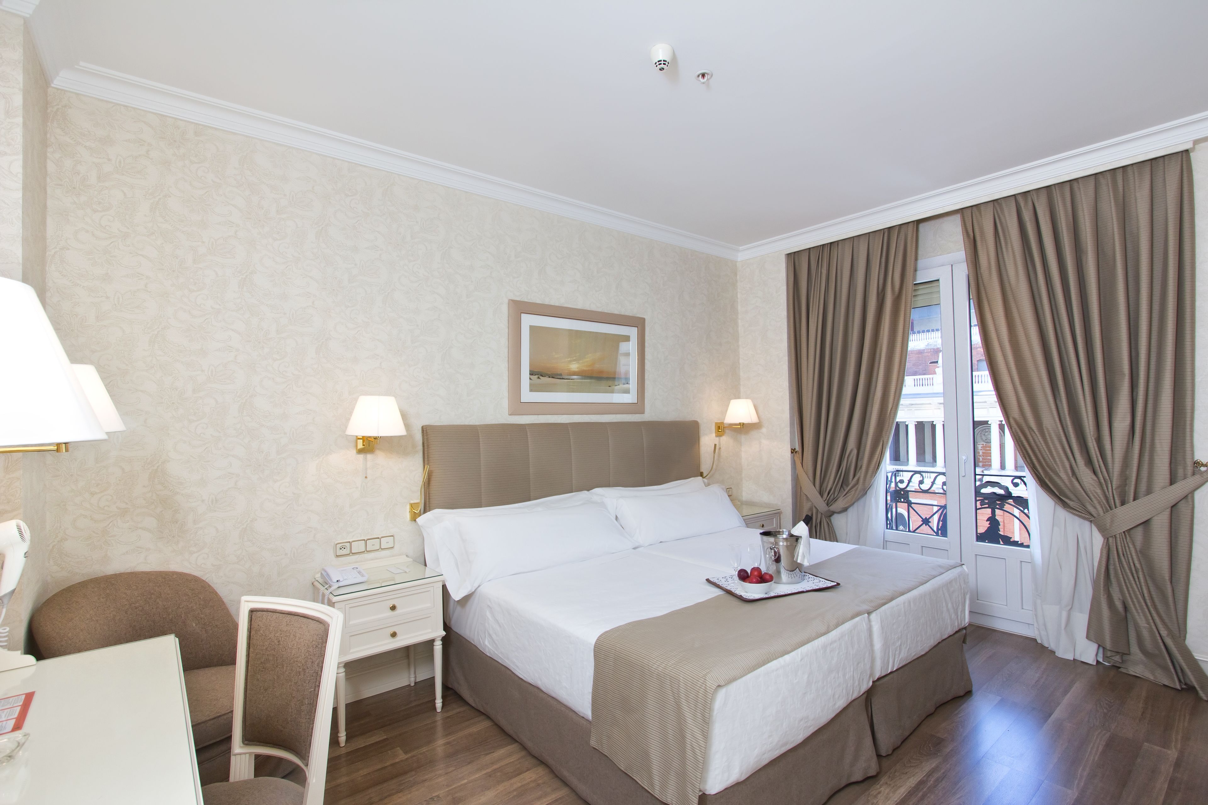 A hotel room with a bed, a desk, and a chair. There is a window with curtains and a view of a balcony.