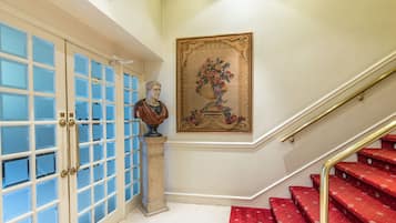 Una escalera con peldaños cubiertos de moqueta roja y barandilla dorada, que conduce a una puerta con paneles de vidrio y una escultura de busto sobre un pedestal.