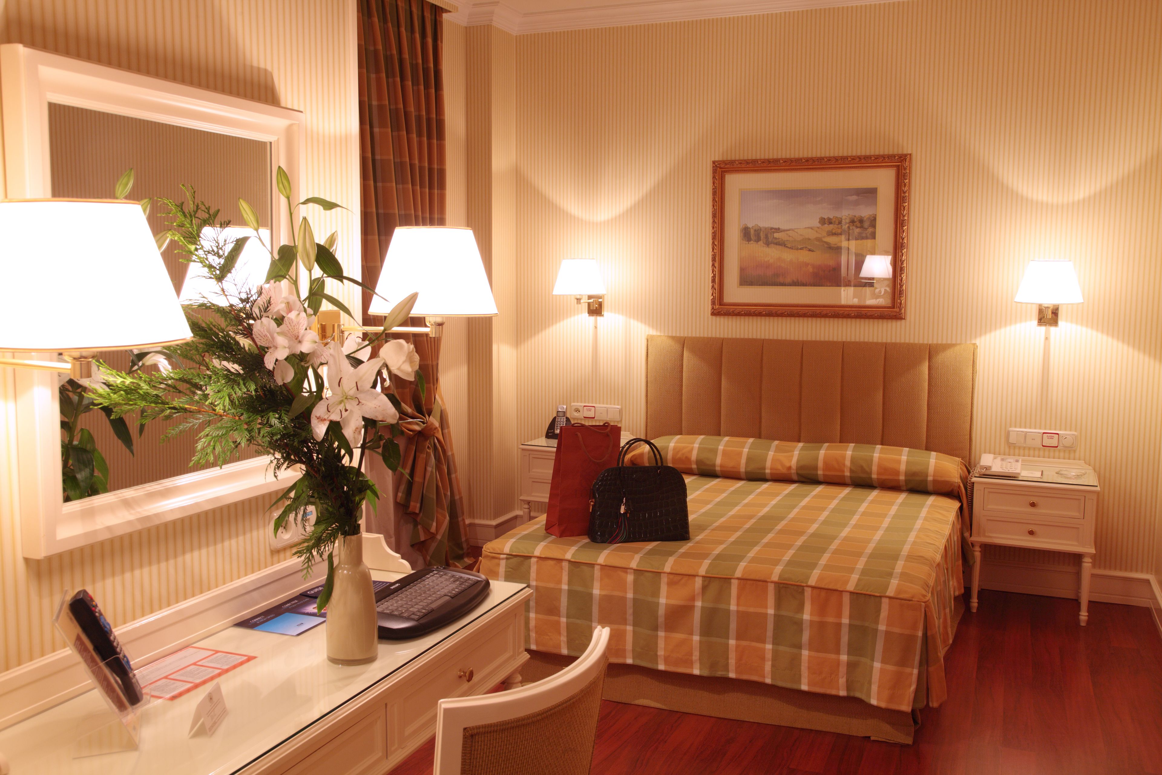 Una camera d'albergo con un letto, una scrivania con un computer, una sedia, una lampada da scrivania, un vaso con fiori, una borsa e un quadro appeso al muro.