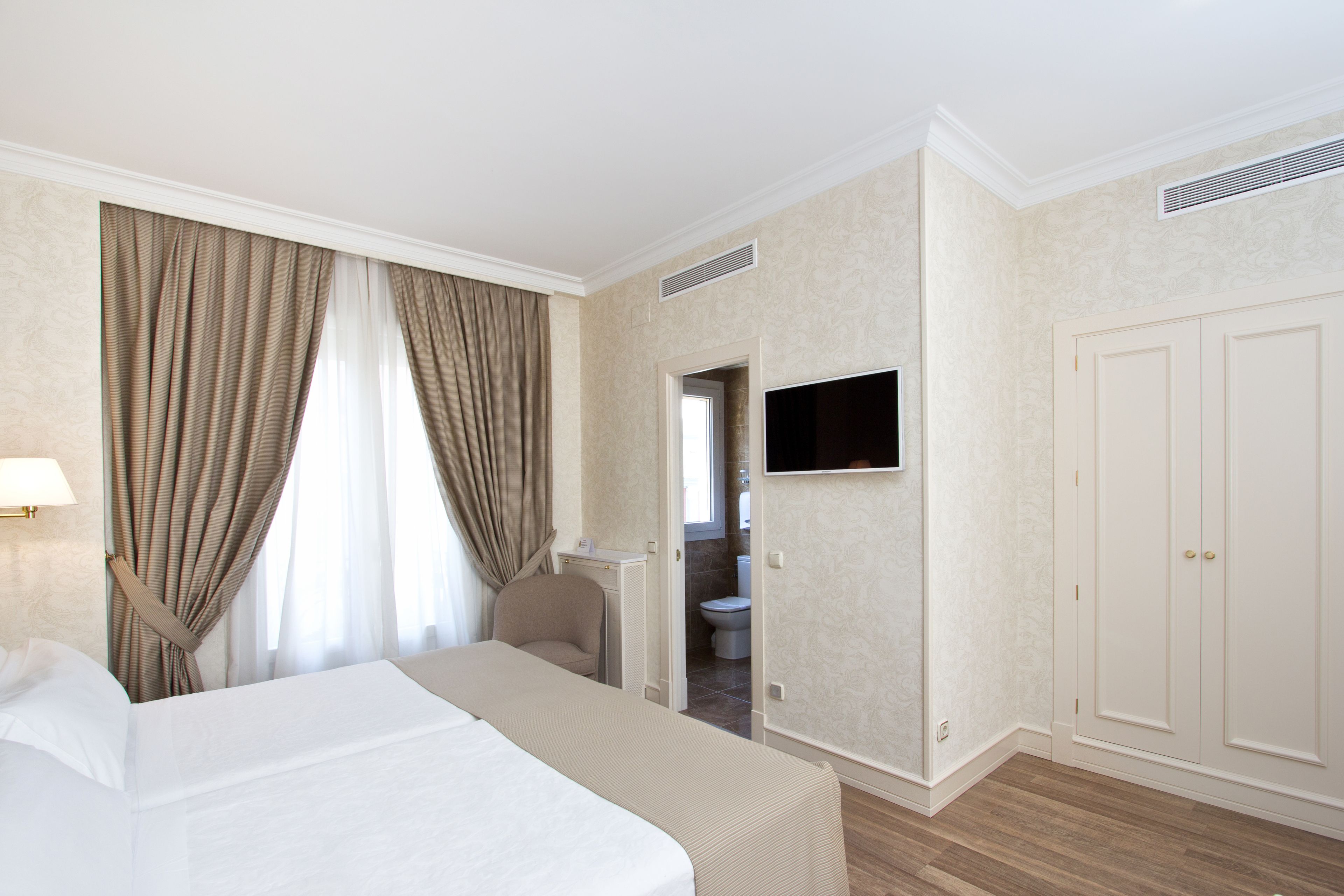 A hotel room with a bed, a TV mounted on the wall, a wardrobe, and a bathroom visible through an open door.