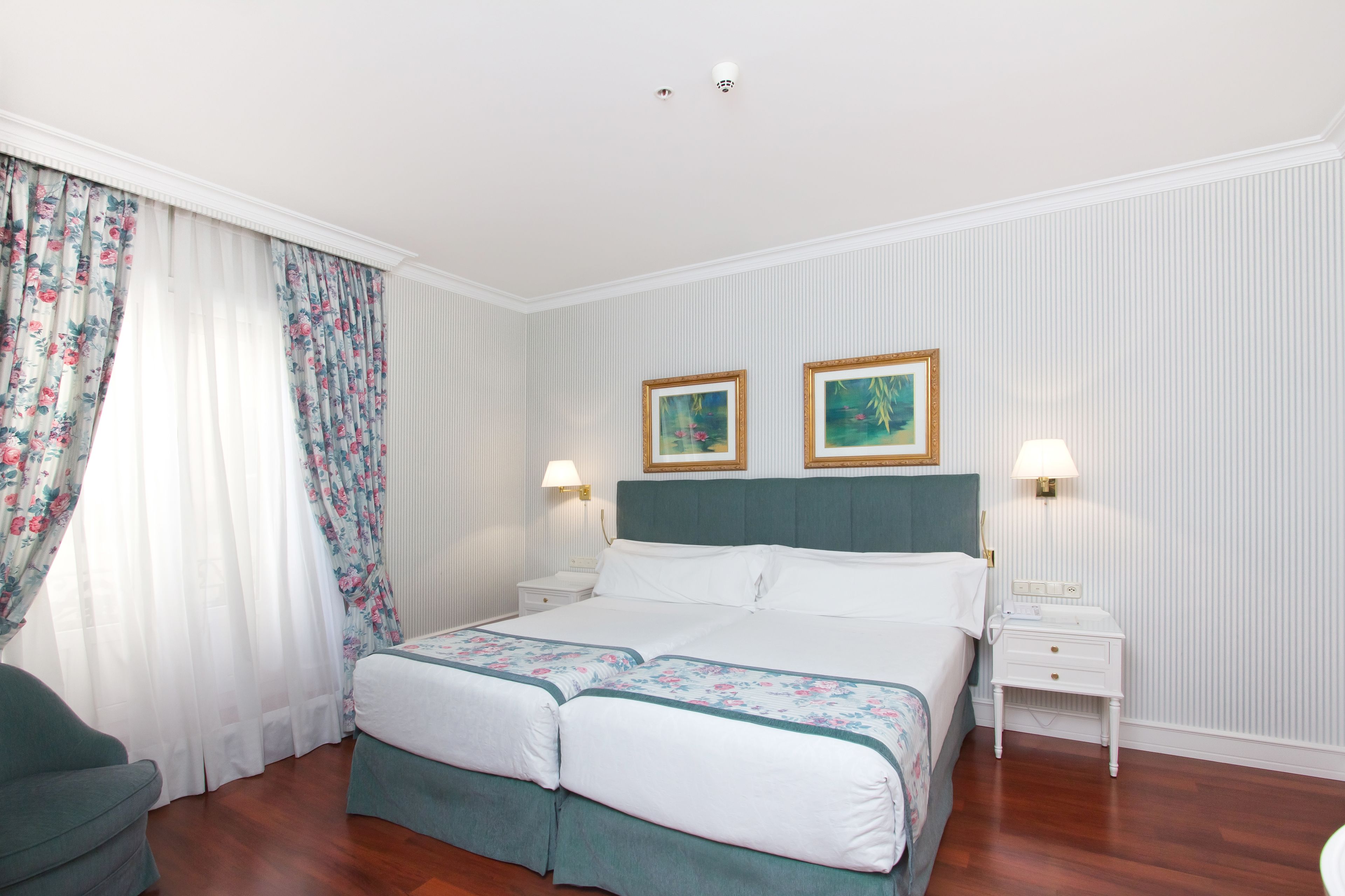 Un letto matrimoniale con testiera verde, due comodini con lampade, tende floreali e due quadri appesi al muro.