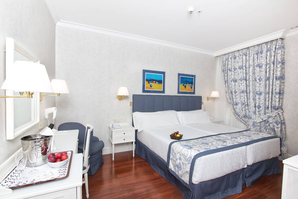 A hotel room with a bed, a desk with a lamp, a chair, a nightstand, and a wall with two framed pictures.
