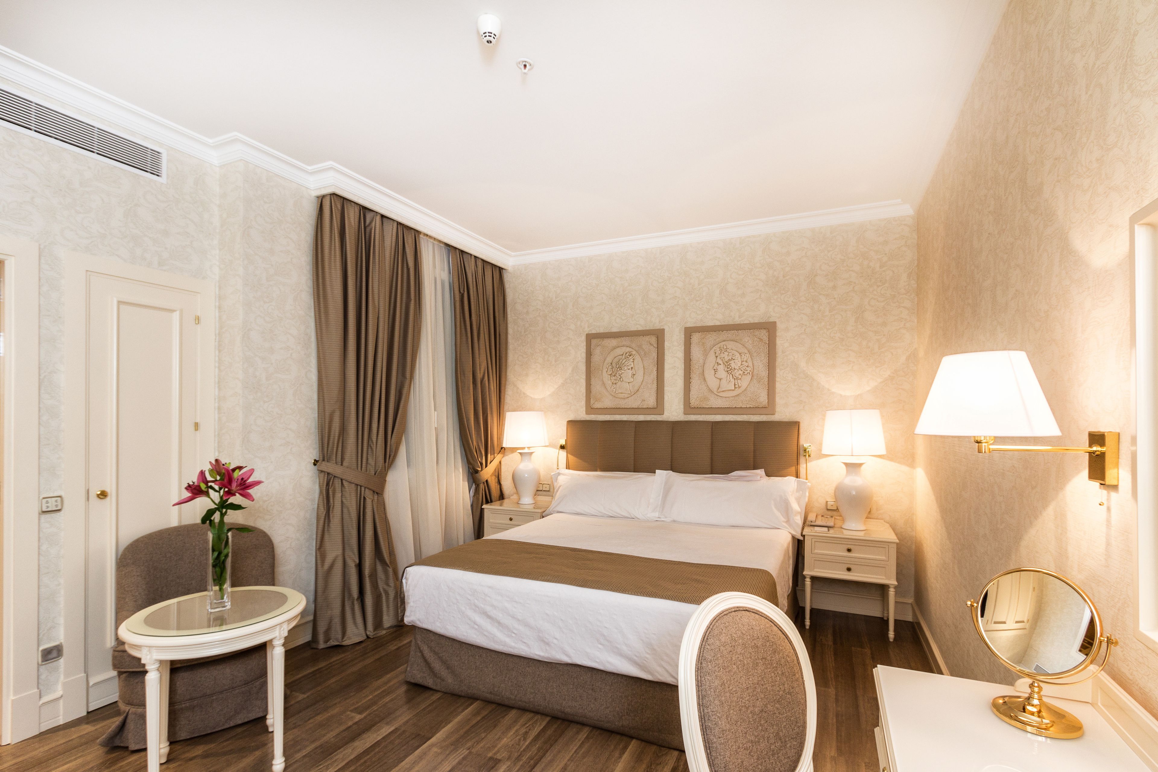 Habitación de hotel con cama, mesitas de noche, una silla, una pequeña mesa con un florero y un espejo.