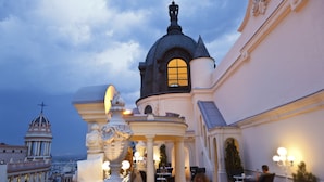 An outdoor terrace with tables and chairs, a decorative fountain, and a view of a cityscape with historical buildings.