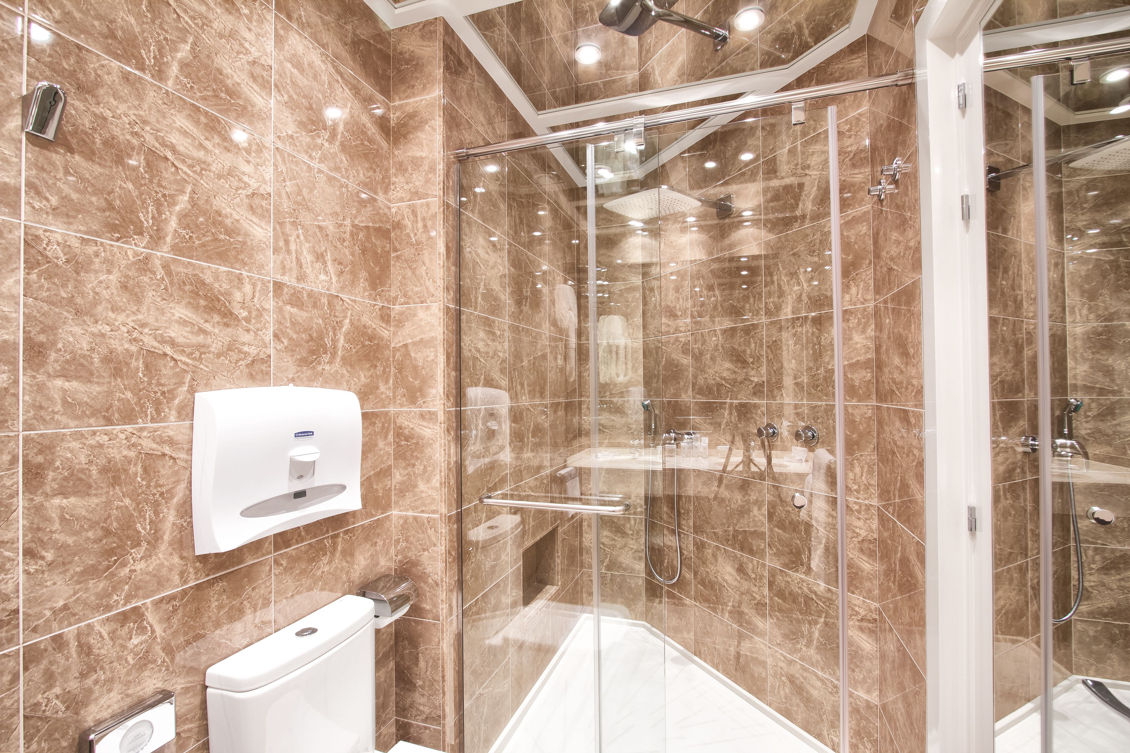A modern bathroom with a glass-enclosed shower, marble tiles, a handheld showerhead, and a soap dispenser.