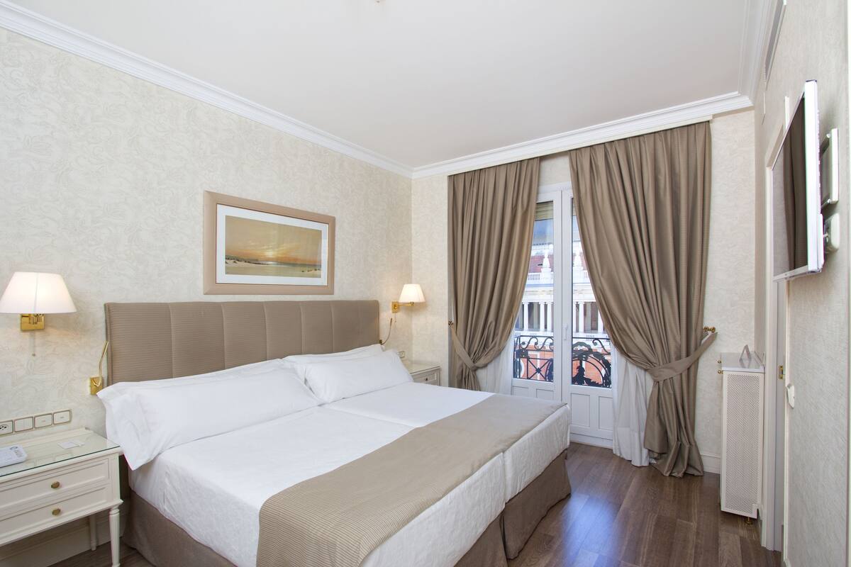 A hotel room with a large bed, two bedside tables with lamps, a framed picture on the wall, and a view of the outside balcony through the window.