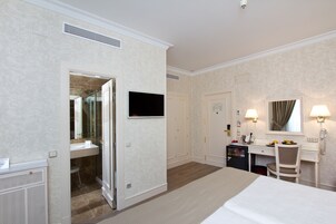 A hotel room with a bed, a desk with a chair, a television, a mirror, and a bathroom with a sink and shower.
