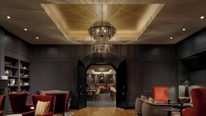 A spacious hotel lobby with a central wooden table, red armchairs, and a chandelier.