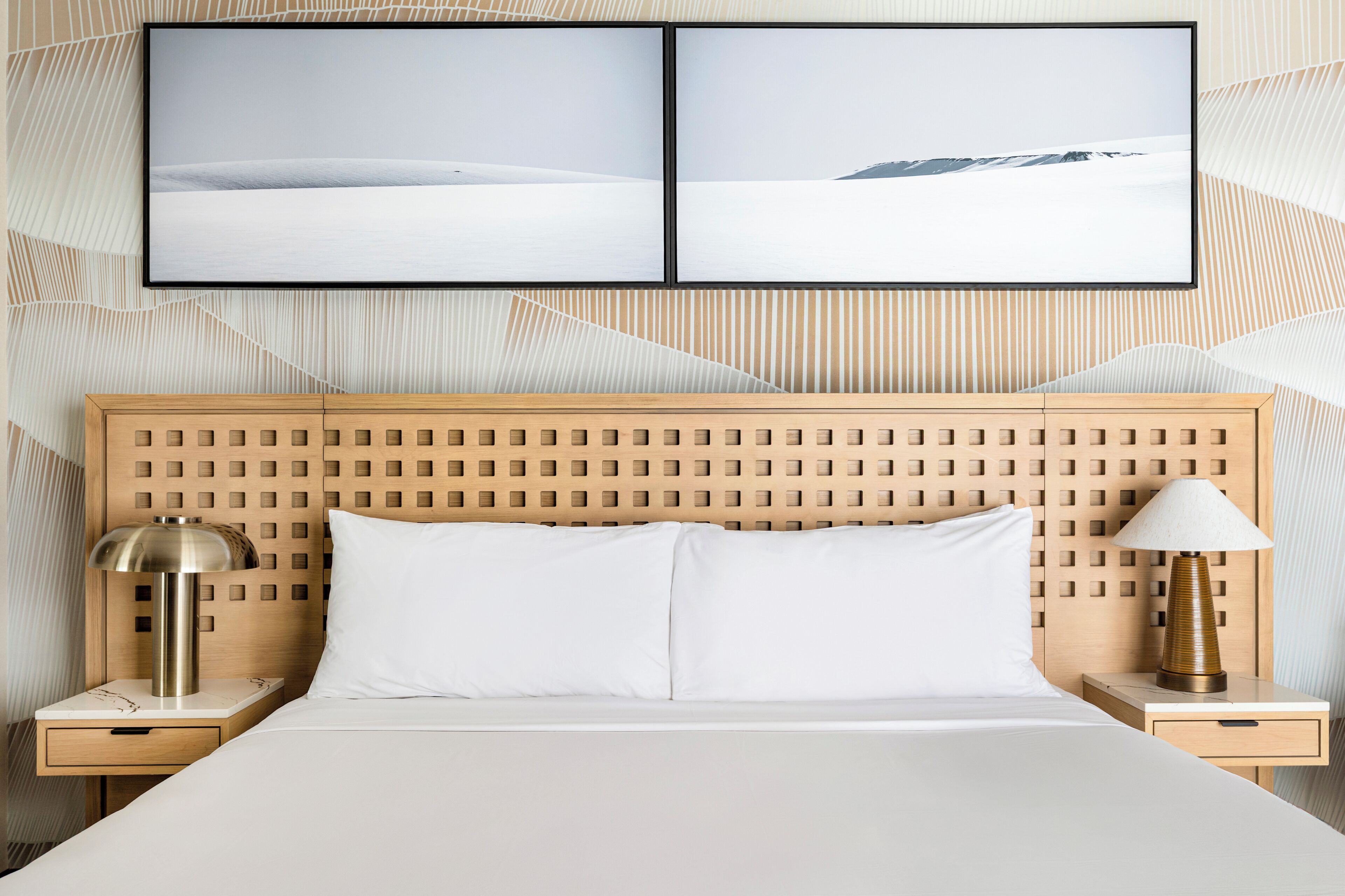 Habitación de hotel con cabecera de madera, dos mesitas de noche, cama con ropa de cama blanca, una lámpara y dos cuadros en la pared.