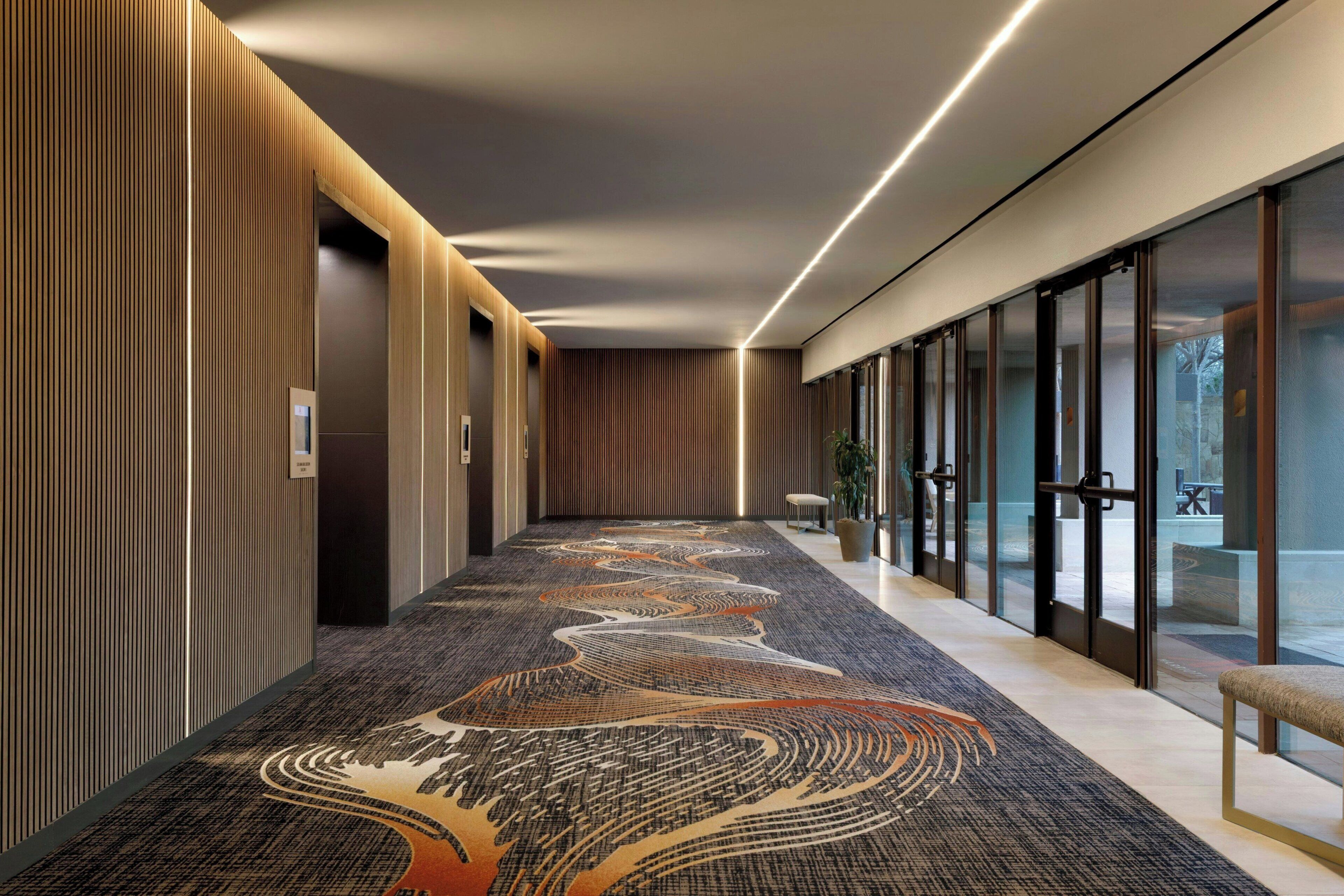 Un couloir d’hôtel moderne, avec un tapis à motifs, des murs recouverts de panneaux en bois et un banc.