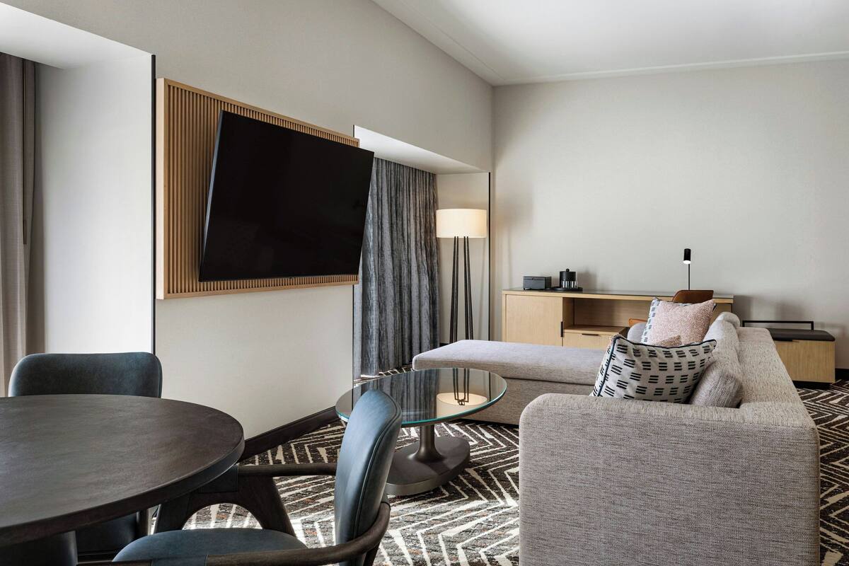 A hotel room with a round dining table, two chairs, a sofa, a TV mounted on the wall, a lamp, and a small cabinet.