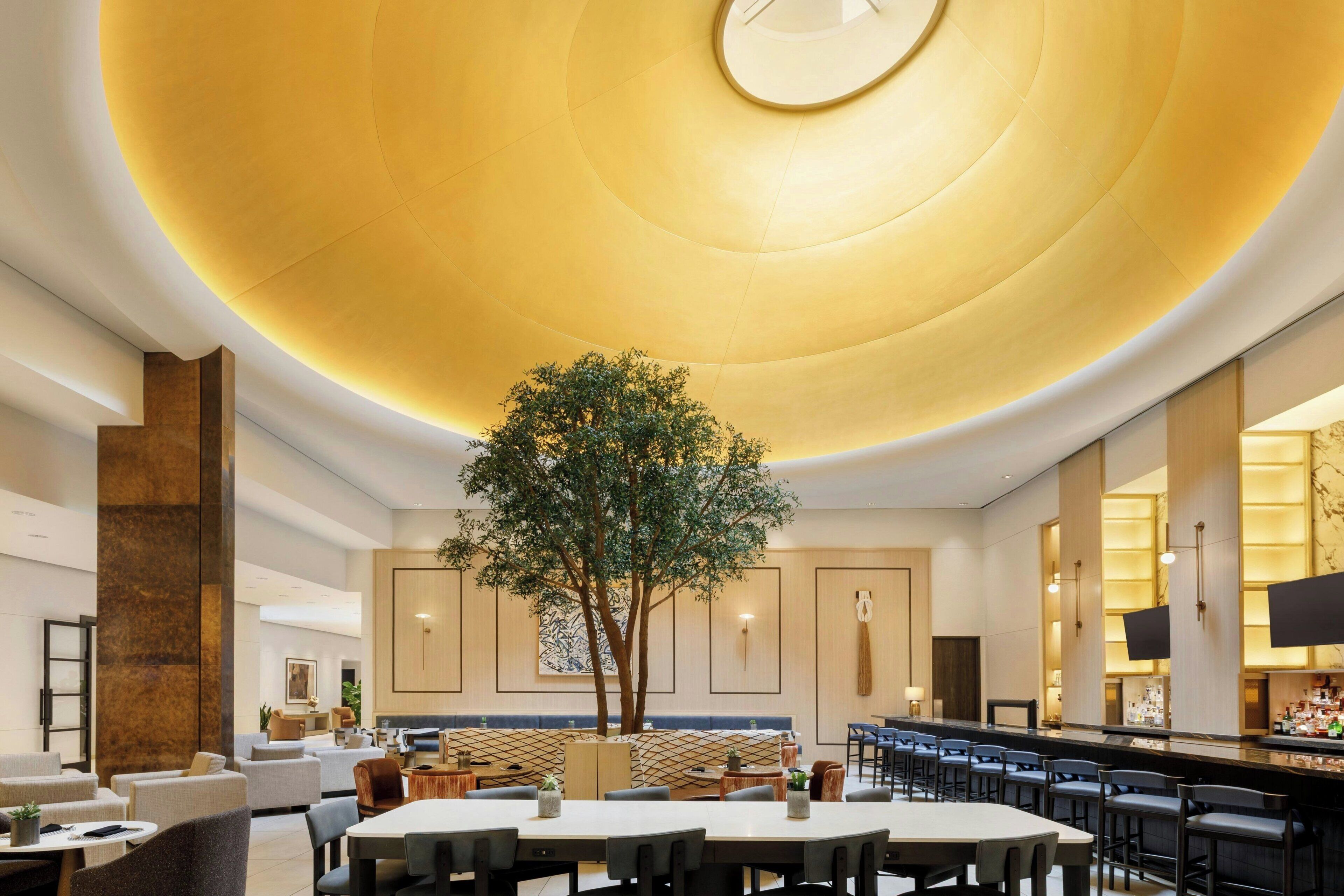 En moderne hotellobby med et stort træ, siddepladser og en bar.