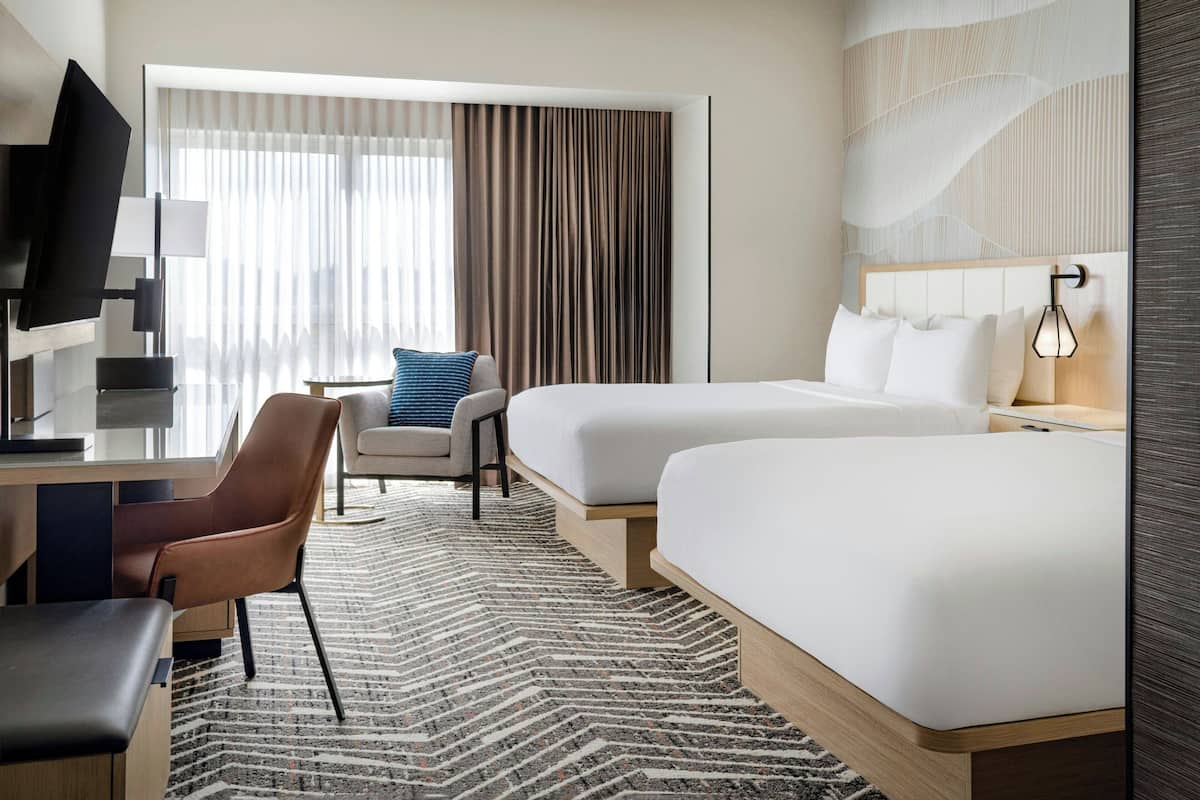 A hotel room with two beds, a desk, a chair, a television, and a large window with curtains.