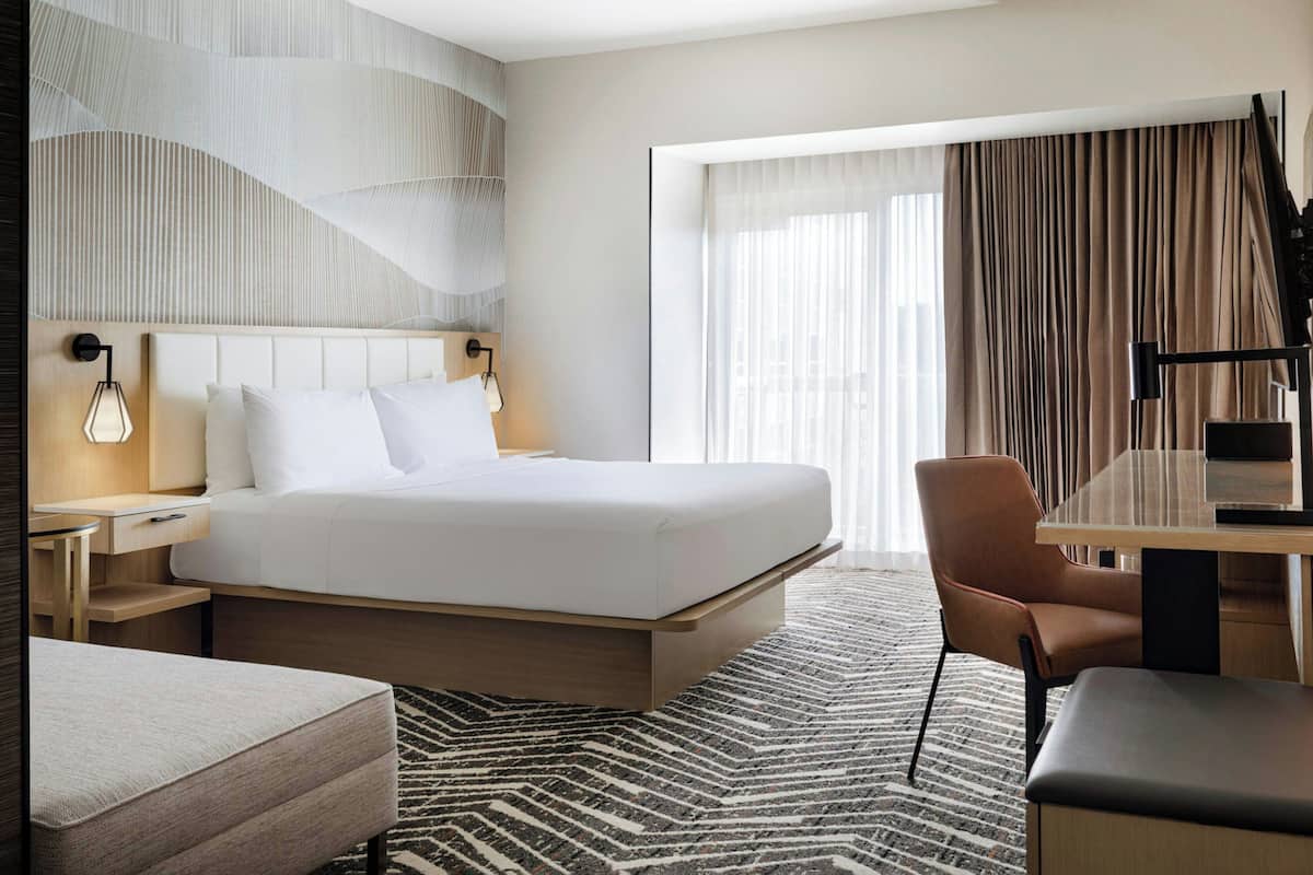 A modern hotel room with a large bed, a desk with a chair, a small sofa, and a large window with curtains.