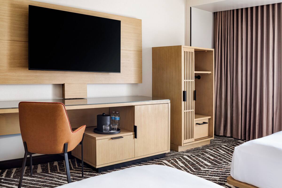 A hotel room with a flat-screen TV mounted on the wall, a wooden desk with a chair, a wardrobe, a bed, and a patterned rug.