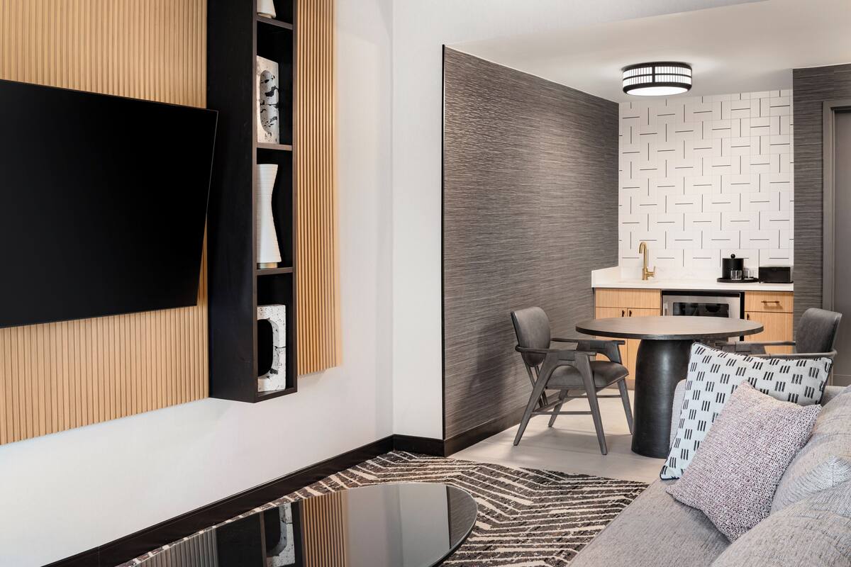 A modern living room with a flat-screen TV, a dining area with a round table, and a kitchen with a patterned backsplash.
