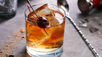 A glass of amber-colored beverage with ice cubes and a cherry on a skewer, placed on a wooden surface with a metal shaker and a glass in the background.