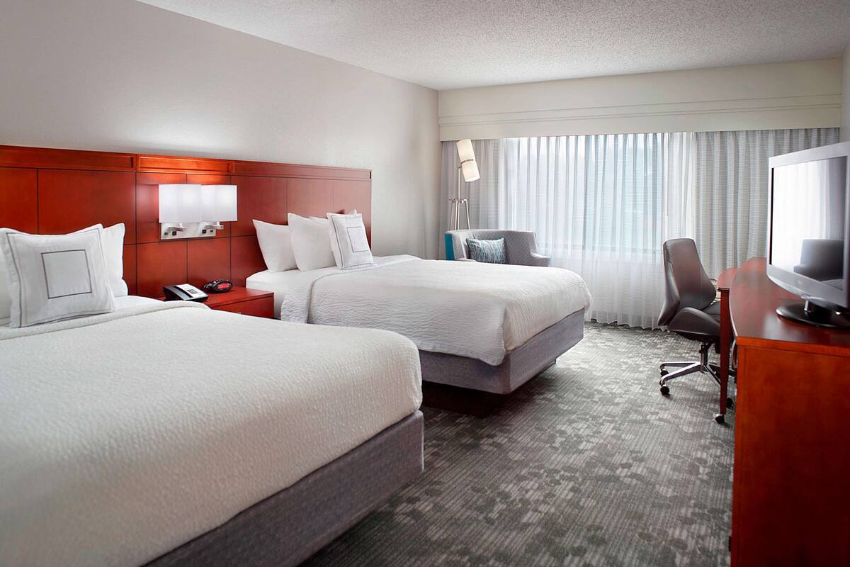 A hotel room with two beds, a desk, a chair, a TV, and a window with curtains.