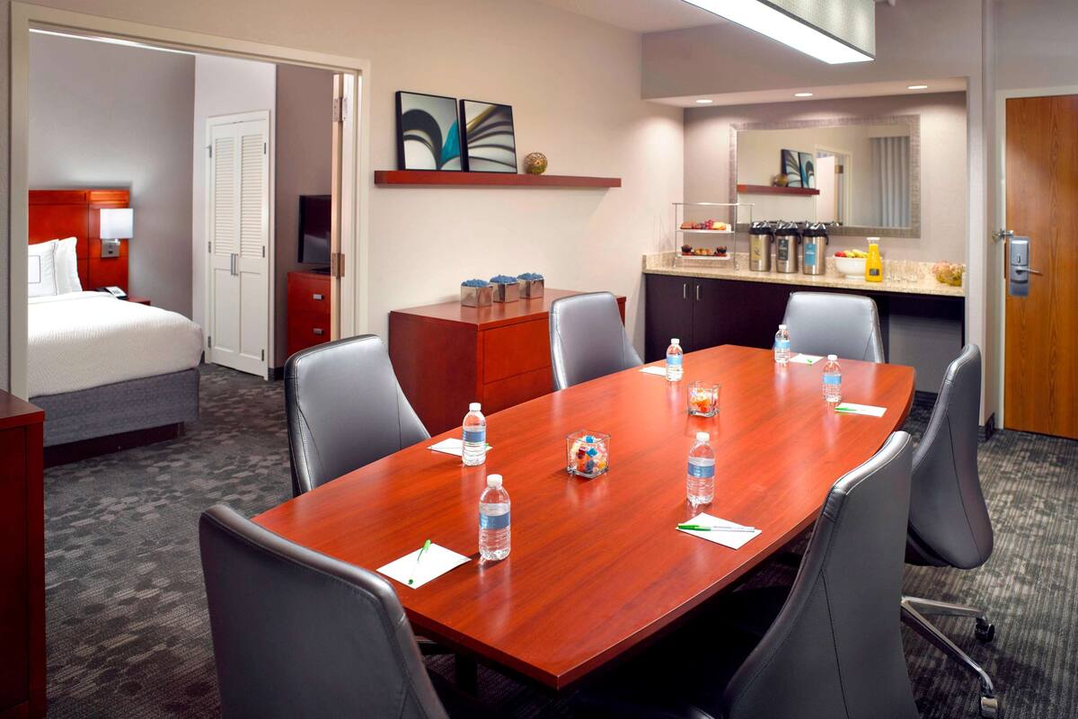 A hotel room with a conference table, chairs, a bed, a TV, a minibar, and a bathroom visible through an open door.