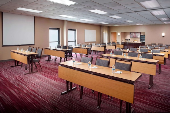 A conference room with multiple tables and chairs, a projector screen, and a kitchen area in the background. - Sonesta Select Atlanta Airport North (Hapeville)