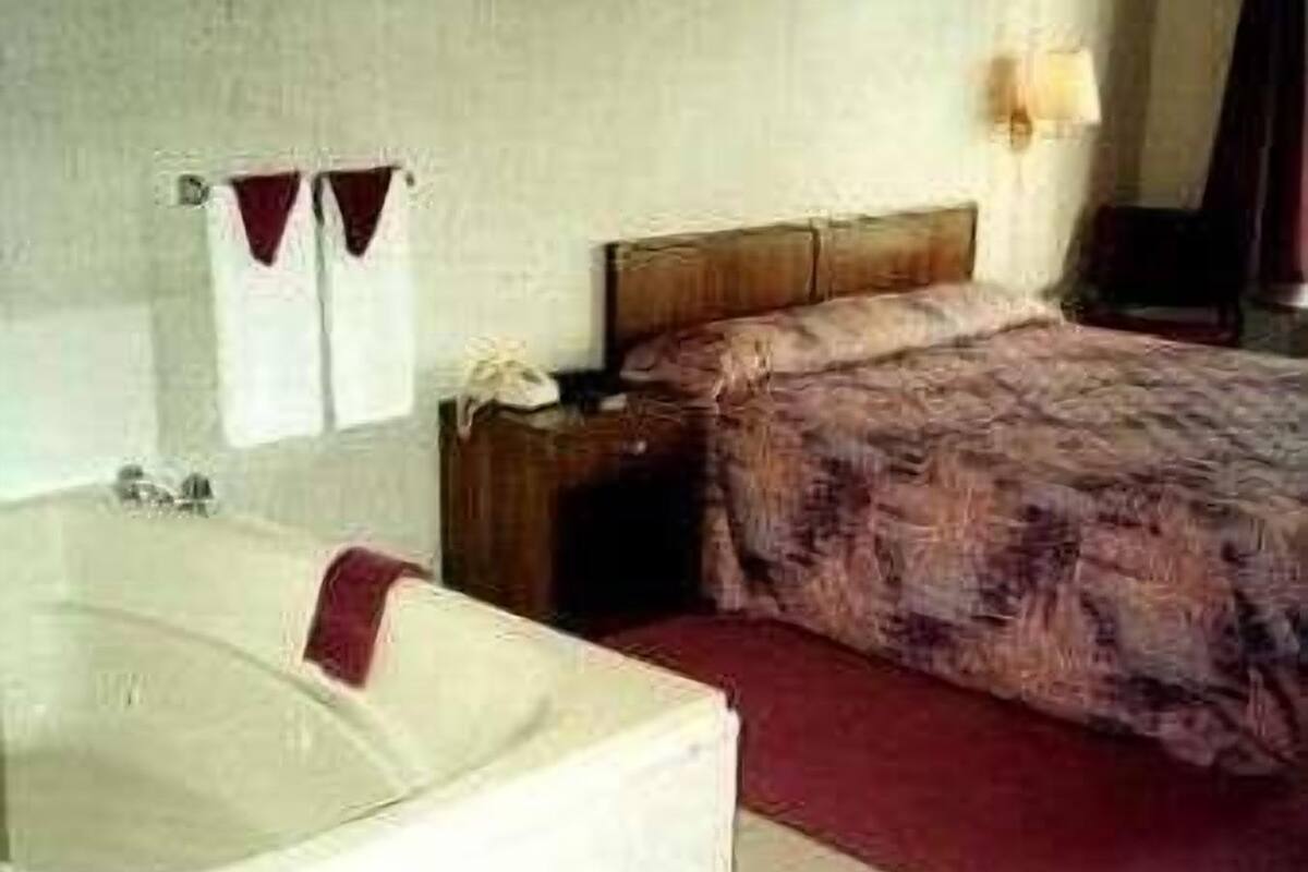 Habitación de hotel con una bañera grande, una mesita de noche de madera, una cama con colcha estampada y una lámpara de pared.