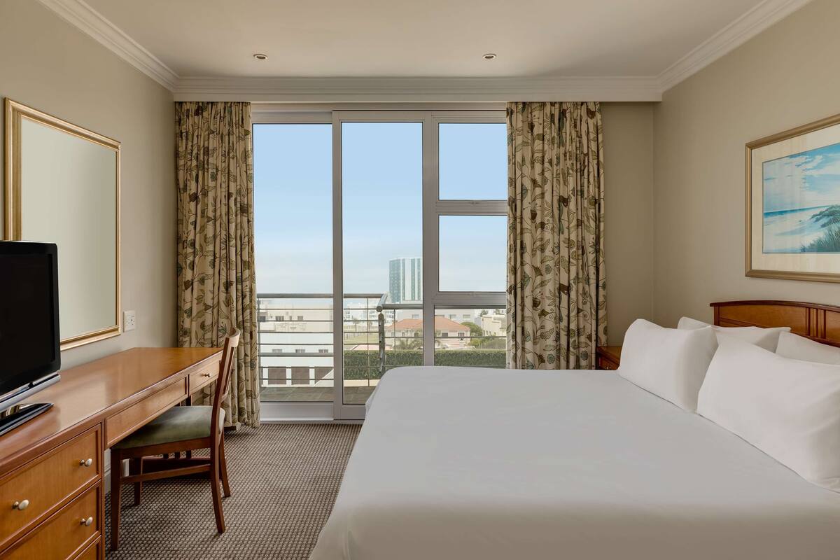 A hotel room with a large bed, a desk with a chair, a television, and a balcony with curtains.