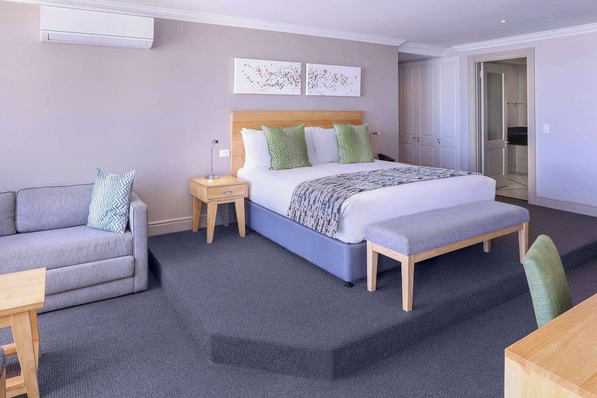 A modern hotel room with a bed, a grey sofa, a wooden nightstand, and a bench.