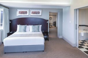 Quarto de hotel com cama grande, cabeceira, duas imagens emolduradas na parede, mesa de cabeceira e banheiro visível através de uma porta aberta.