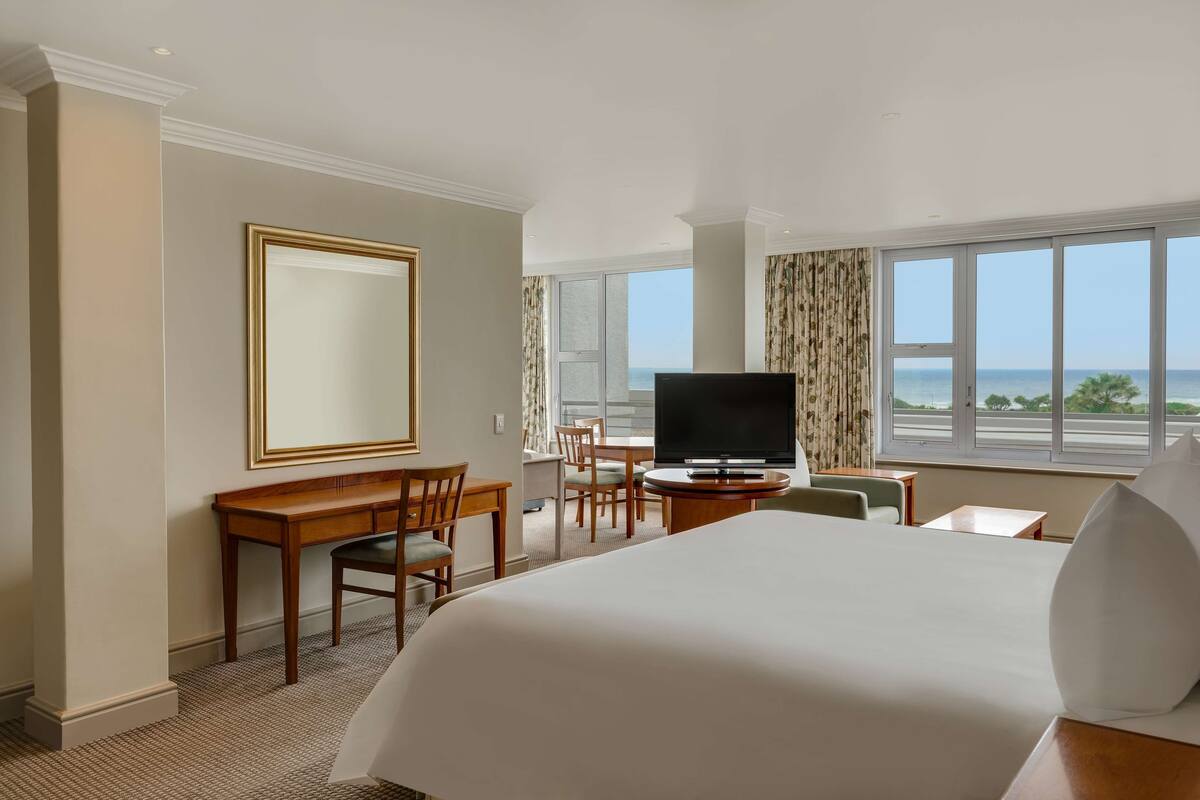 A hotel room with a large bed, a desk with a chair, a television, and a view of the ocean through large windows.