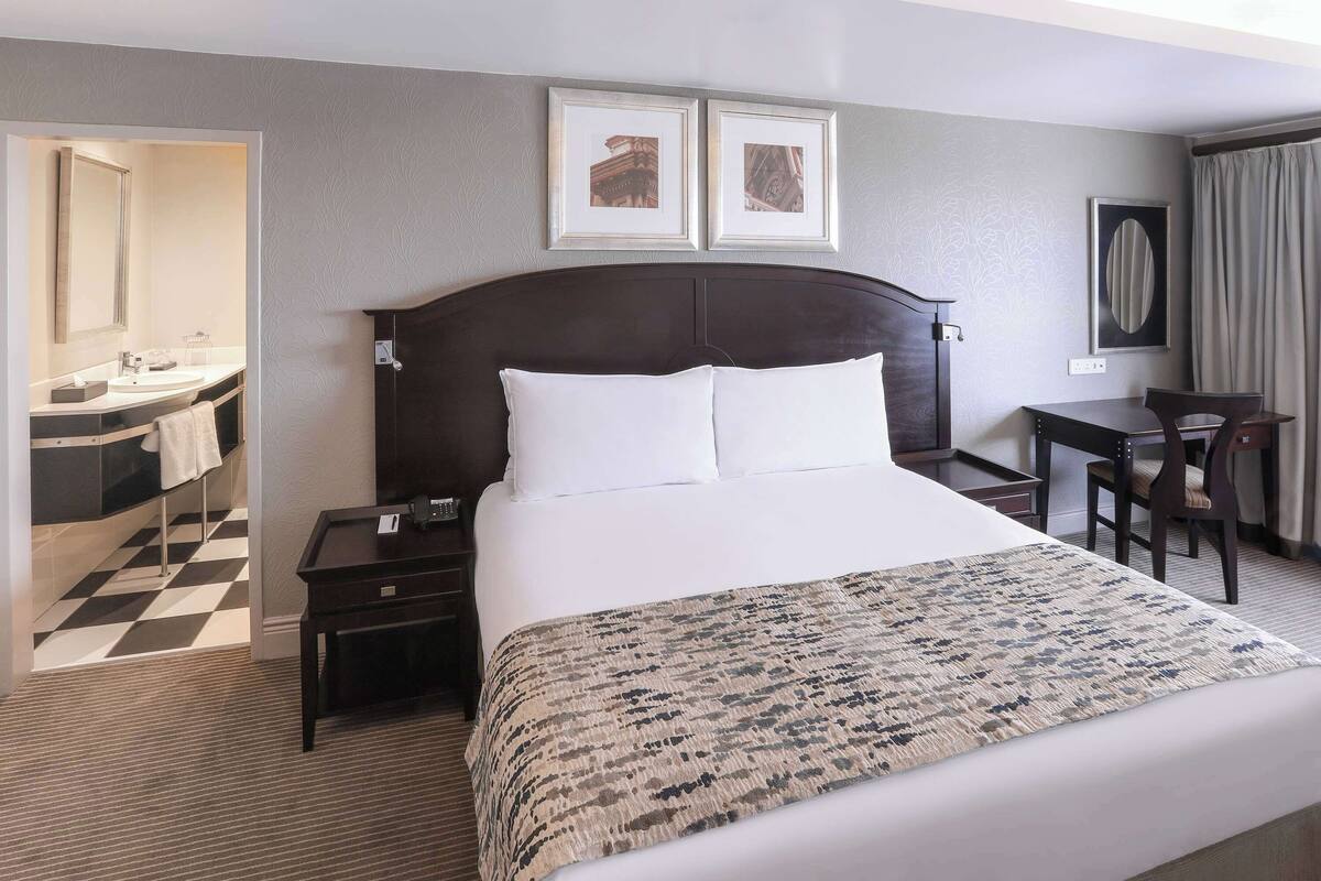 A hotel room with a large bed, a desk, a chair, a mirror, and a bathroom with a checkered floor.