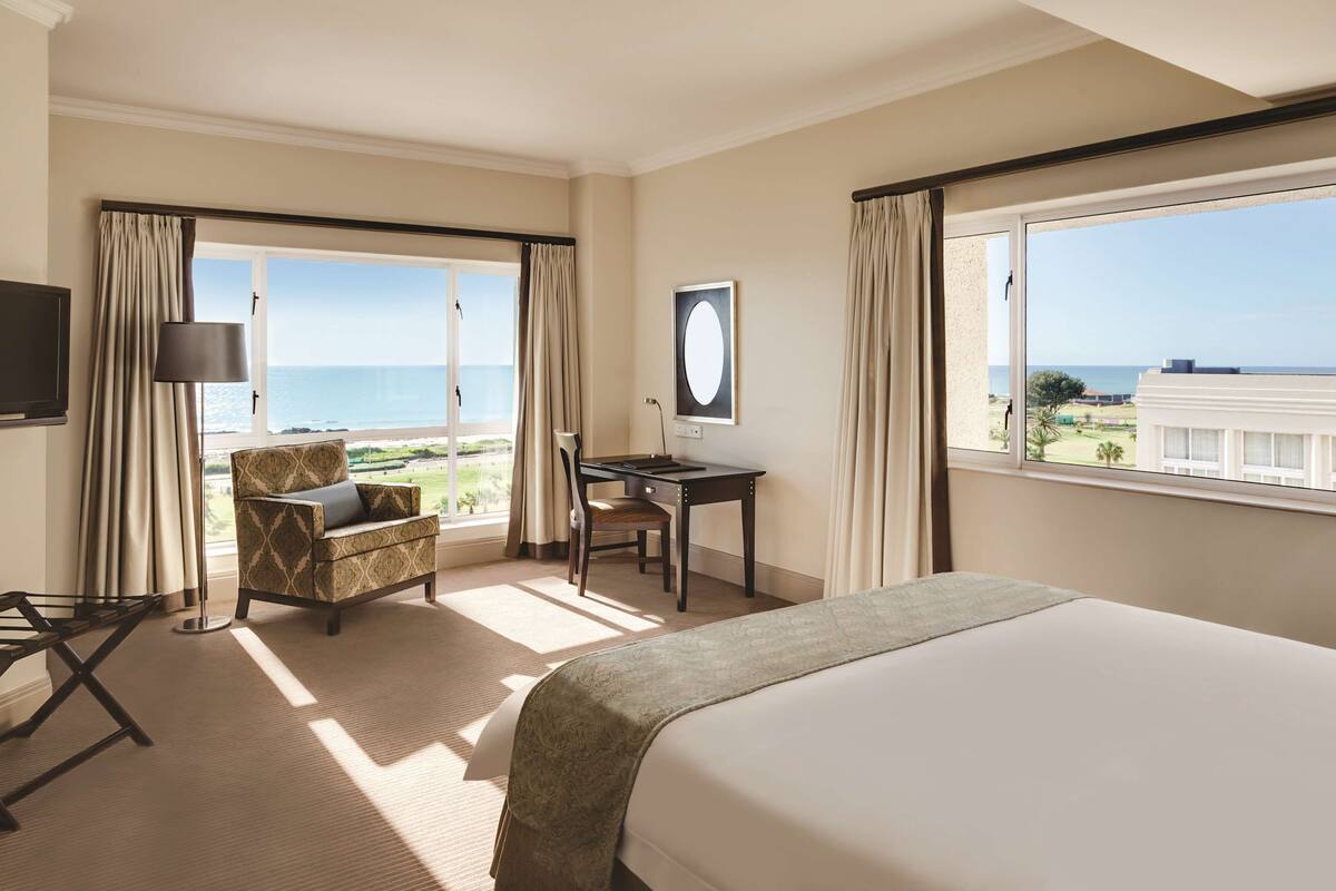 A hotel room with a bed, a desk, a chair, and a TV. There are large windows with curtains, and a view of the ocean.