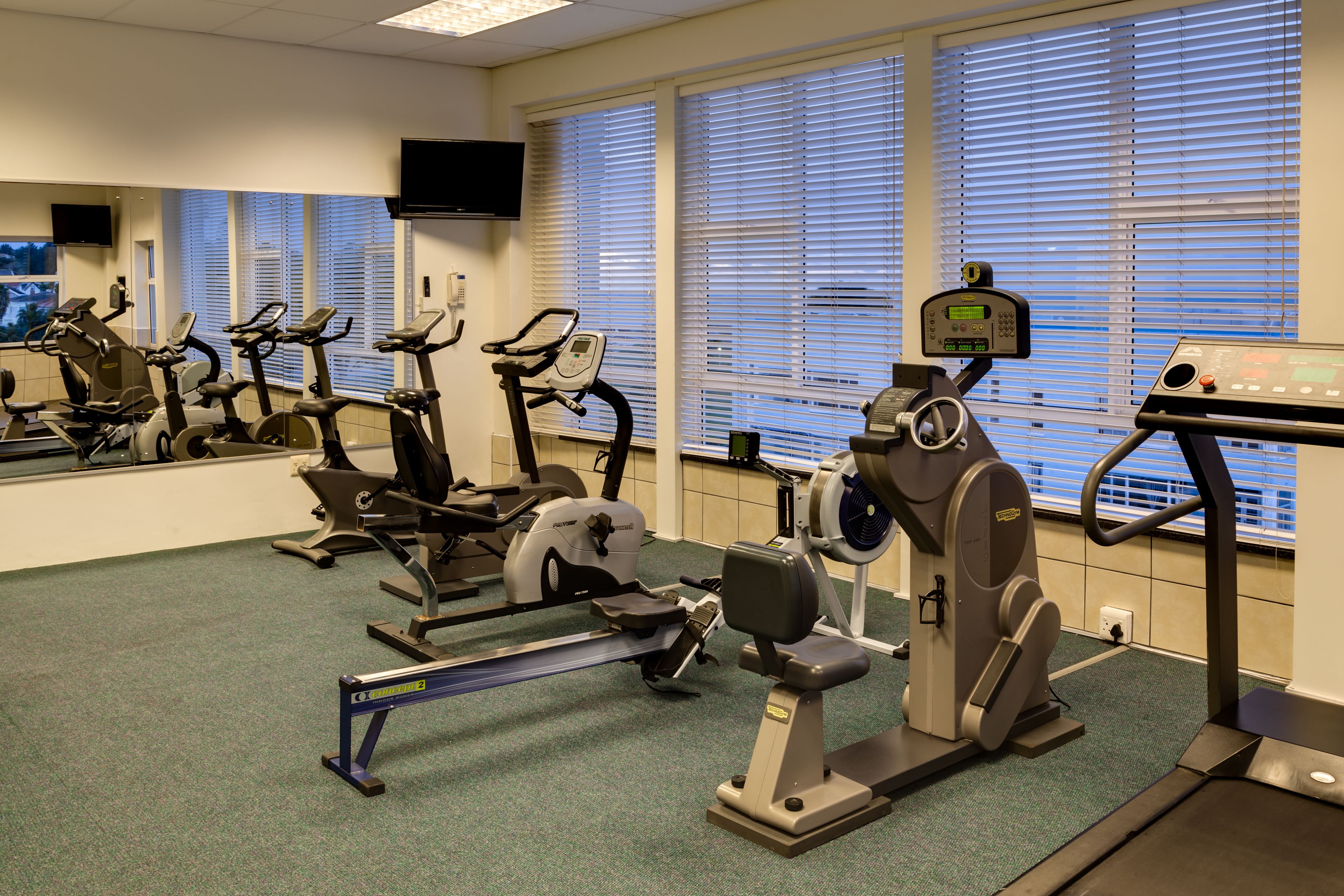 A gym with various exercise equipment, including treadmills and stationary bikes, and large windows with blinds.