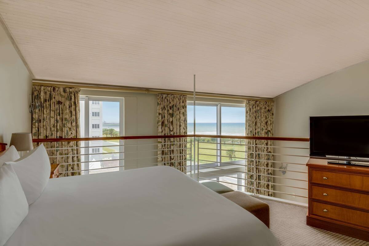 A hotel room with a large bed, a TV, a wooden dresser, and a balcony with a view of the city.