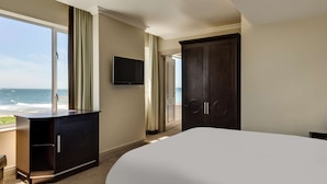 Une chambre d’hôtel avec un grand lit, une armoire en bois sombre, un téléviseur fixé au mur et une vue sur l’océan par une grande fenêtre.