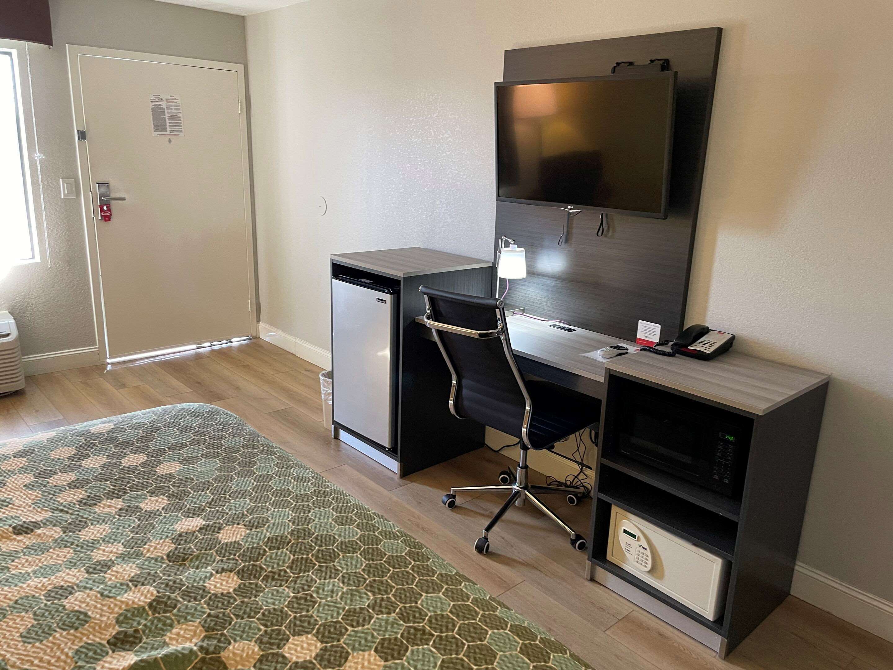 Habitación de hotel compacta con cama, escritorio con computadora, televisión y refrigerador pequeño.