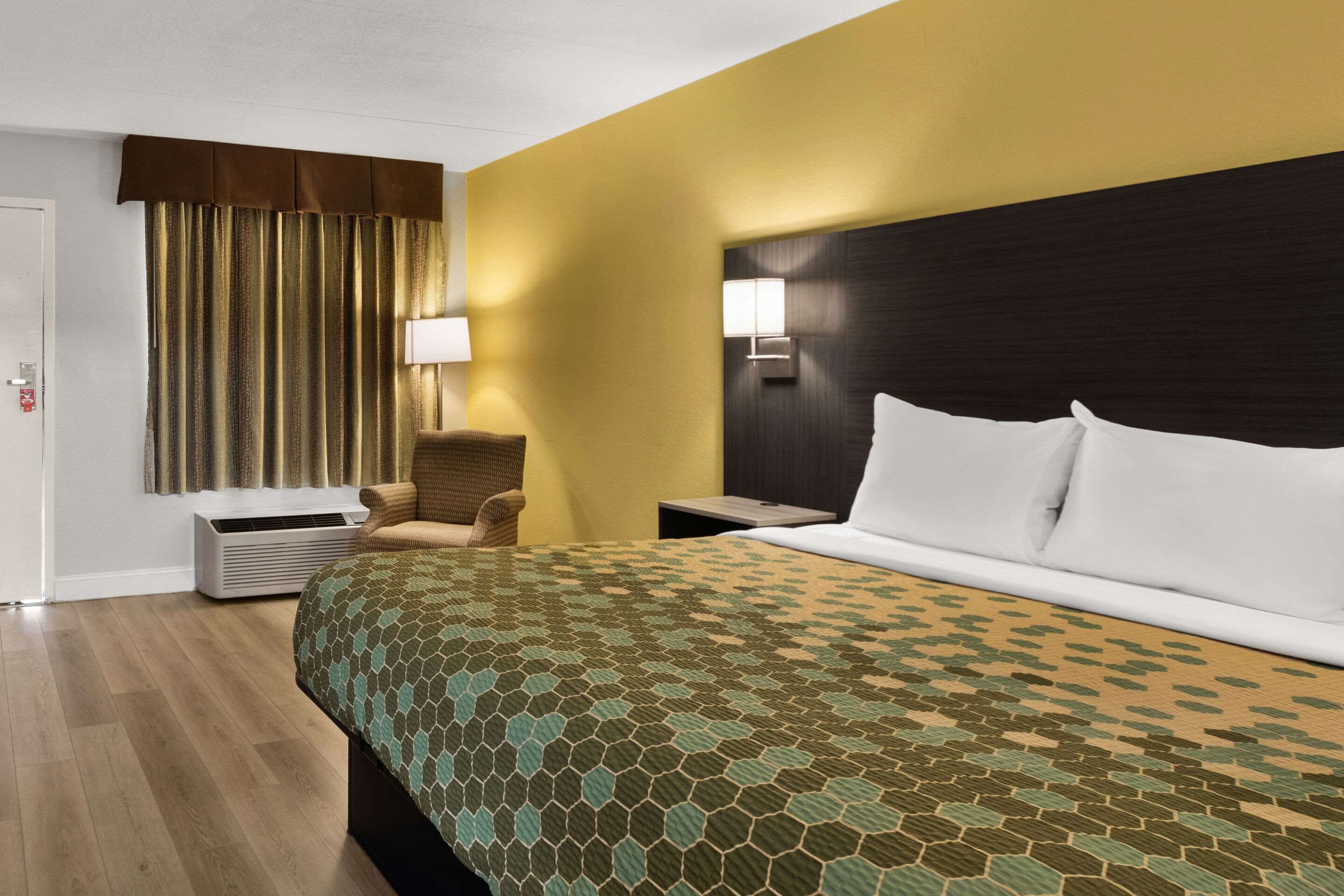 A hotel room with a bed, a chair, a nightstand, a lamp, and a wall-mounted air conditioner.