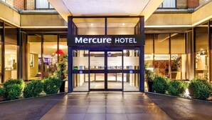 Der Eingang zum Mercure Hotel mit Glasfassade, einem Schild über den Türen und einem mit Fliesen belegten Weg, der zum Eingang führt.
